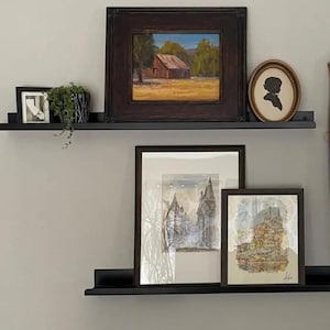 Two black floating mrtal shelves with various framed artwork. The top shelf has a framed landscape painting with a brown wooden frame, a small black framed photo, and a small potted plant. The middle shelf has a framed black and white drawing with a white mat and a brown wooden frame. The bottom shelf has a framed watercolor painting with a white mat and a brown wooden frame.