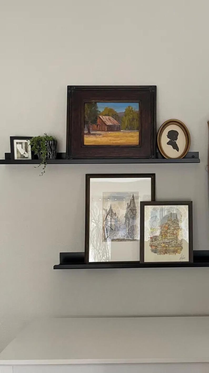 Two black floating mrtal shelves with various framed artwork. The top shelf has a framed landscape painting with a brown wooden frame, a small black framed photo, and a small potted plant. The middle shelf has a framed black and white drawing with a white mat and a brown wooden frame. The bottom shelf has a framed watercolor painting with a white mat and a brown wooden frame.