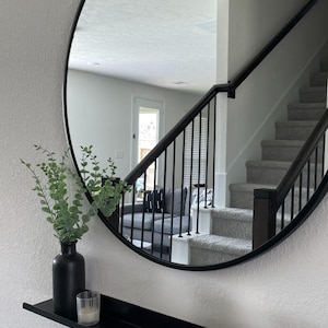 A black round mirror with a floating black metal shelf underneath. The mirror reflects a staircase with a black railing and a white wall. A small green plant is in a black vase on the shelf.