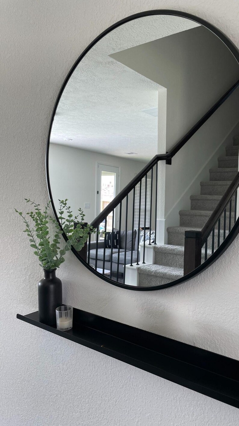 A black round mirror with a floating black metal shelf underneath. The mirror reflects a staircase with a black railing and a white wall. A small green plant is in a black vase on the shelf.