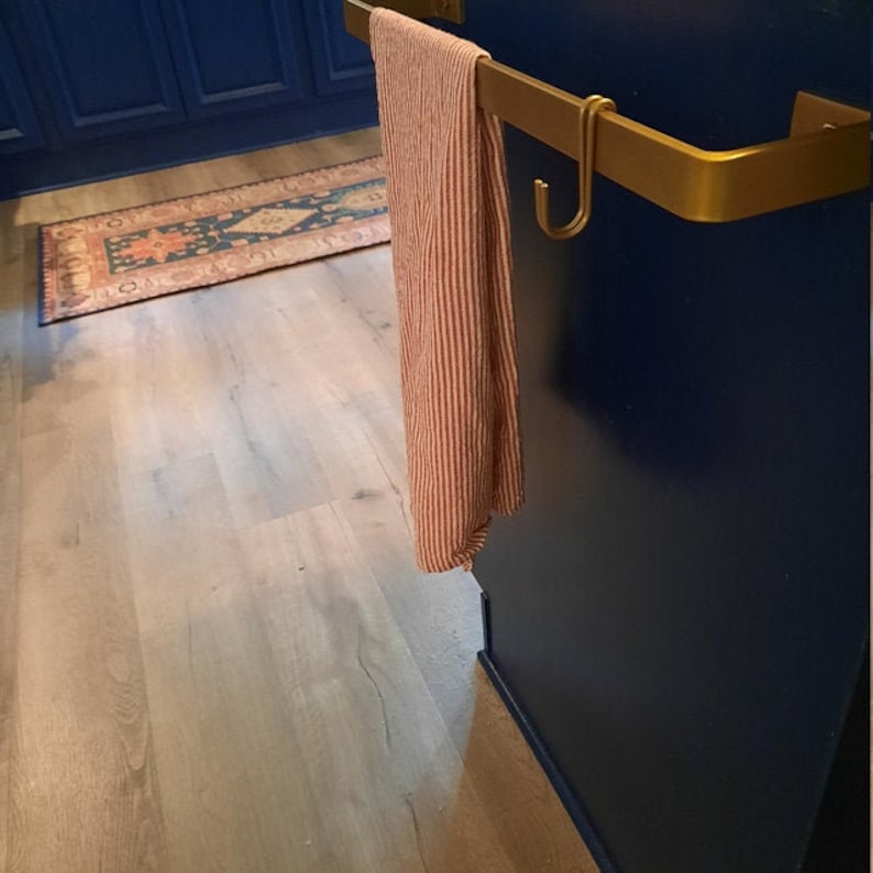 Navy kitchen cabinet with gold towel bar with s hooks for kitchen mounted on the side. There is a towel on the hook rack and a rug in the background.