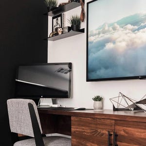 A modern home office with a floating desk, a black computer monitor, a gray office chair, and a framed print of clouds. The desk has two drawers with black handles. There are two black metal shelves mounted on the wall with various decorative items.