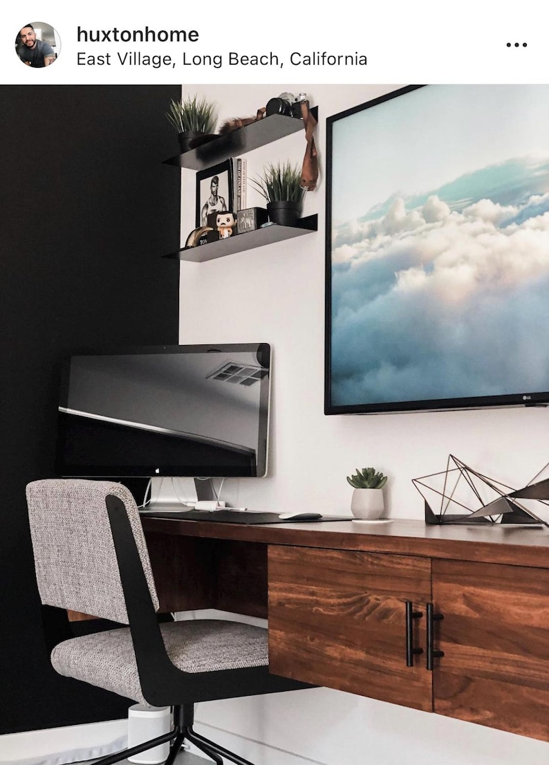 A modern home office with a floating desk, a black computer monitor, a gray office chair, and a framed print of clouds. The desk has two drawers with black handles. There are two black metal shelves mounted on the wall with various decorative items.
