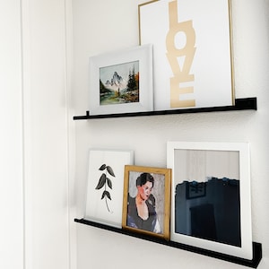 May include: A black floating shelf with four framed pictures. The top shelf has a white frame with a landscape painting and a white frame with the word "LOVE" in gold letters. The bottom shelf has a white frame with a black and white leaf drawing, a gold frame with a portrait of a woman, and a white frame with a blue and white abstract painting.