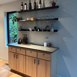 A light wood cabinet with four drawers and two doors. The cabinet has black hardware. Above the cabinet are three black floating shelves with various items on them, including wine bottles, glasses, and coffee equipment.