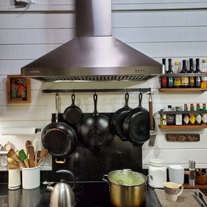 Kitchen with white walls and stove range with metal rack holding pots and pans with hooks. There are also spices and utensils.
