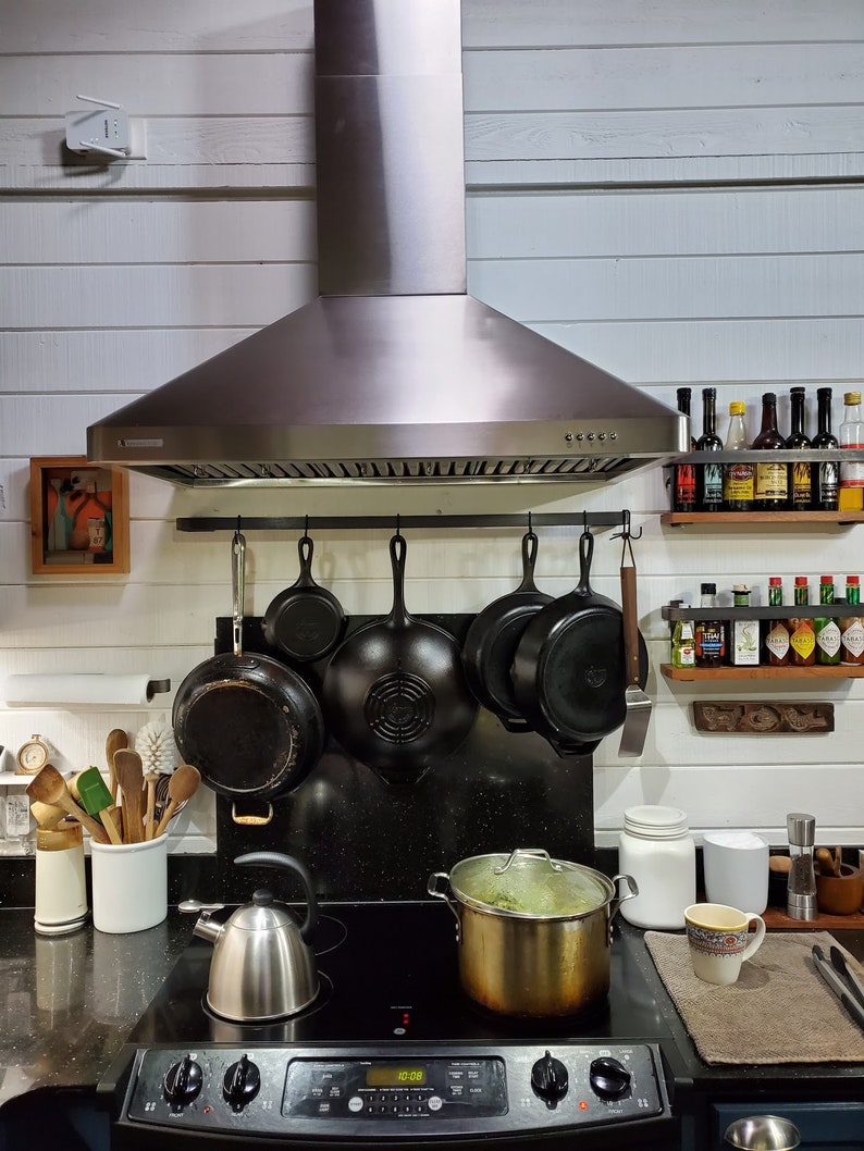 Kitchen with white walls and stove range with metal rack holding pots and pans with hooks. There are also spices and utensils.