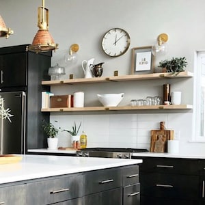 A modern kitchen with black cabinets and a white countertop. There are two wooden shelves with brass brackets, a clock, and a sign that says "Be Kind".
