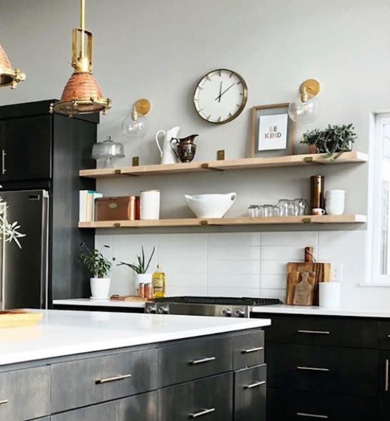 A modern kitchen with black cabinets and a white countertop. There are two wooden shelves with brass brackets, a clock, and a sign that says "Be Kind".