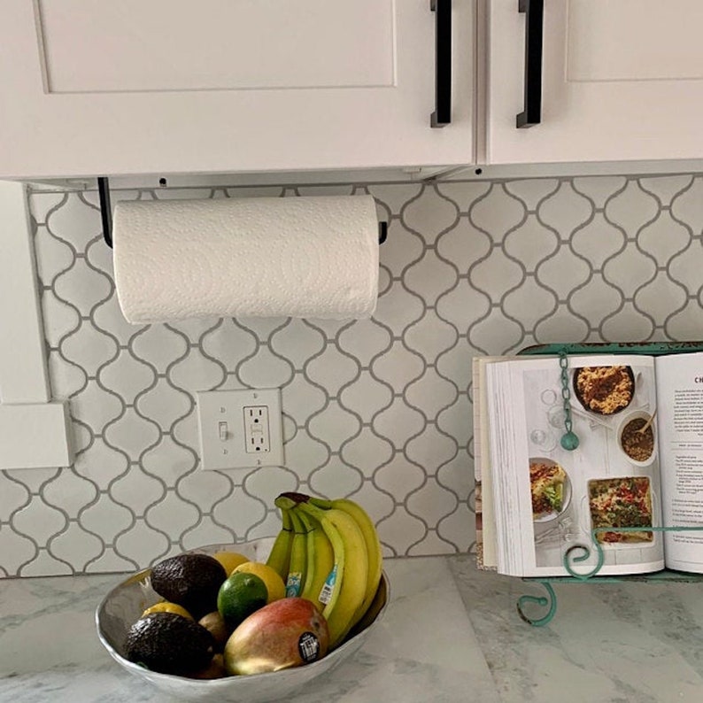 A white bright kitchen with a black paper towel holder mounted under white kitchen cabinets. Light gray countertops and tile backsplash.