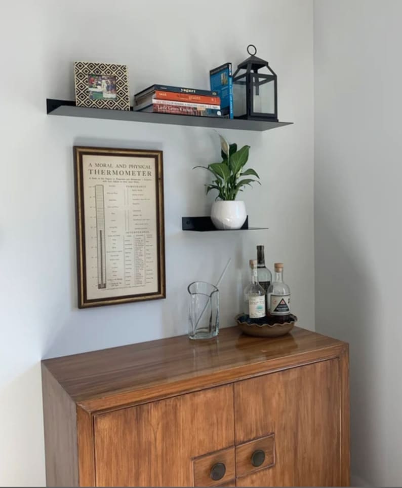 There is a wood cabinet with two floating metal shelves above. The black shelves are mounted on a gray wall and holding books, plants, and decor.