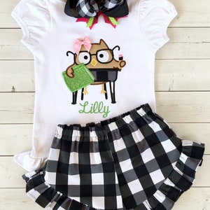 May include: A white short-sleeved shirt with a black and white checkered bow, a green book, and an owl with glasses and a pencil. The shirt has the name "Lilly" embroidered on it. The shirt is paired with black and white checkered shorts with a ruffled hem.