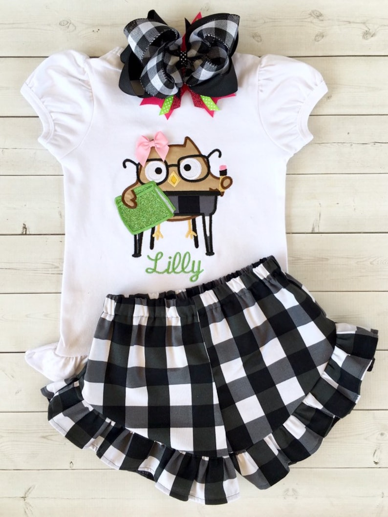 May include: A white short-sleeved shirt with a black and white checkered bow, a green book, and an owl with glasses and a pencil. The shirt has the name "Lilly" embroidered on it. The shirt is paired with black and white checkered shorts with a ruffled hem.
