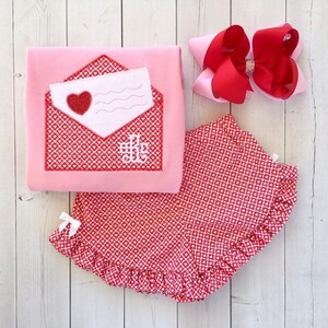 May include: A pink and red Valentine's Day outfit for a baby or toddler. The outfit includes a pink shirt with an embroidered red envelope and a red and white heart pattern skirt.