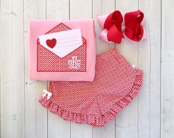 May include: A pink and red Valentine's Day outfit for a baby or toddler. The outfit includes a pink shirt with an embroidered red envelope and a red and white heart pattern skirt.