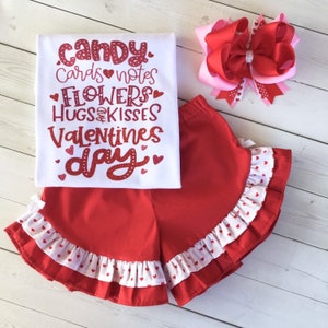May include: Red and white Valentine's Day outfit for a child. The outfit includes a white t-shirt with red glitter text that says "Candy, Cards, Notes, Flowers, Hugs & Kisses, Valentines Day" and red shorts with white ruffle trim and white hearts.