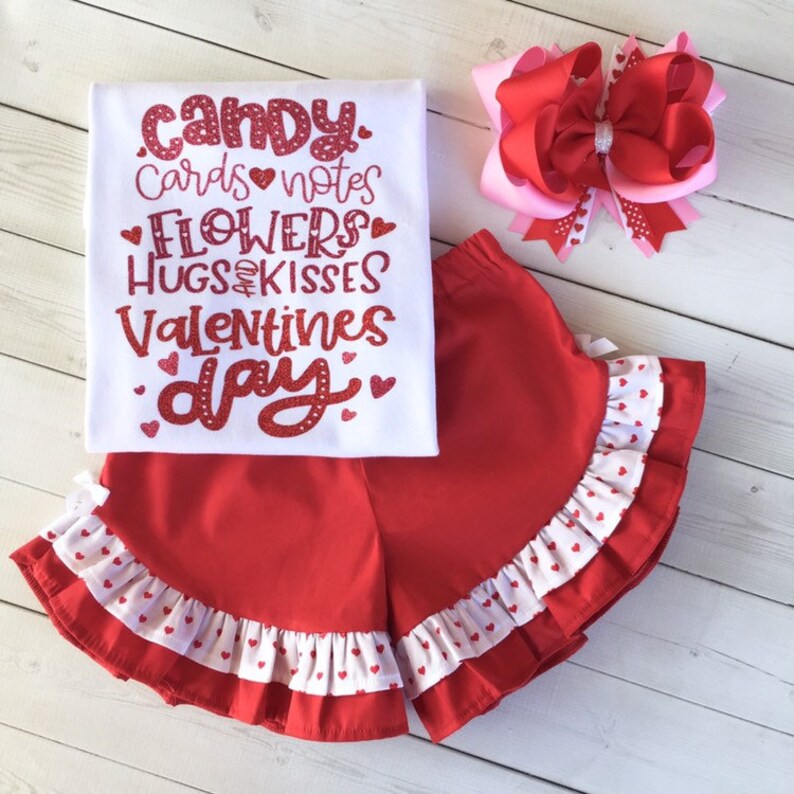 May include: Red and white Valentine's Day outfit for a child. The outfit includes a white t-shirt with red glitter text that says "Candy, Cards, Notes, Flowers, Hugs & Kisses, Valentines Day" and red shorts with white ruffle trim and white hearts.