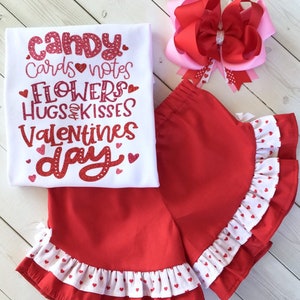 May include: A white t-shirt with red glitter text that says "Candy, Cards, Notes, Flowers, Hugs & Kisses, Valentines Day". A red and white polka dot skirt with a ruffle trim. A pink and red bow.