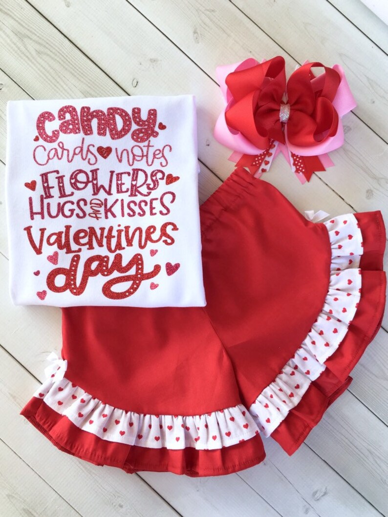May include: A white t-shirt with red glitter text that says "Candy, Cards, Notes, Flowers, Hugs & Kisses, Valentines Day". A red and white polka dot skirt with a ruffle trim. A pink and red bow.
