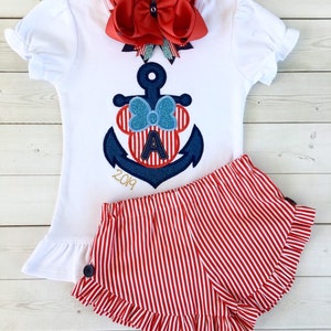 May include: A white short-sleeved shirt with a red and blue anchor and a red bow on top. The anchor has a red and white striped Mickey Mouse head with the letter A in the middle. The shirt also has the year 2019 embroidered on it. The shirt is paired with red and white striped shorts with a ruffle trim.