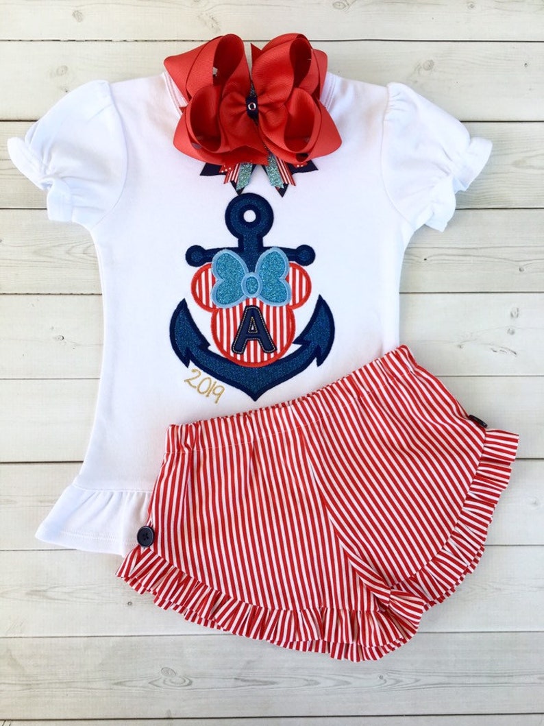 May include: A white short-sleeved shirt with a red and blue anchor and a red bow on top. The anchor has a red and white striped Mickey Mouse head with the letter A in the middle. The shirt also has the year 2019 embroidered on it. The shirt is paired with red and white striped shorts with a ruffle trim.