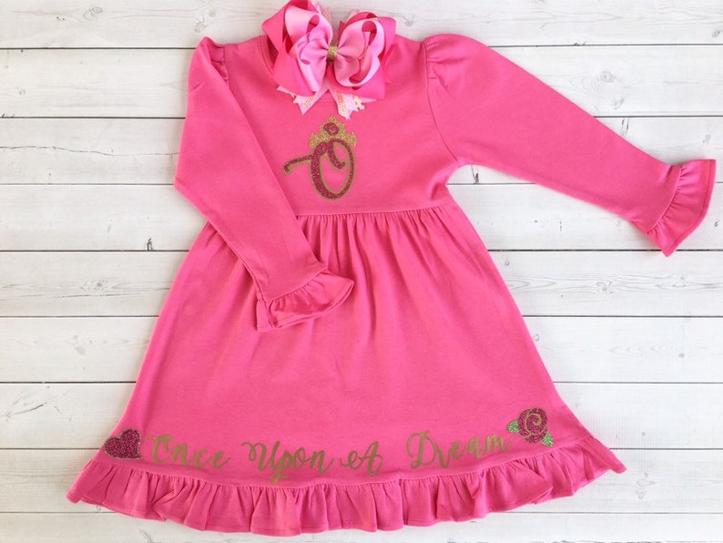 Peut inclure: Robe rose &agrave; manches longues avec une couronne dor&eacute;e paillet&eacute;e et la lettre "O" sur le devant. La robe a un ourlet et des manches &agrave; volants. Le texte "Once Upon A Dream" est &eacute;crit en paillettes dor&eacute;es sur le bas de la robe.