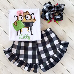May include: A white t-shirt with a brown owl wearing glasses and holding a green book. The shirt has the name "Lilly" embroidered on it. The outfit also includes black and white checkered shorts with a ruffle trim and a black and white checkered hair bow.