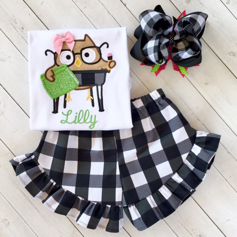 May include: A white t-shirt with a brown owl wearing glasses and holding a green book. The shirt has the name "Lilly" embroidered on it. The outfit also includes black and white checkered shorts with a ruffle trim and a black and white checkered hair bow.