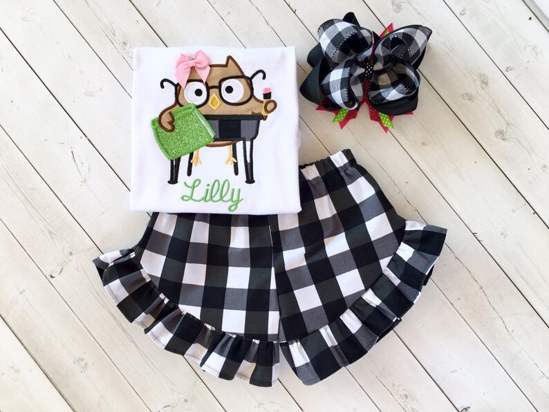 May include: White shirt with a black and brown owl wearing glasses and holding a green book. The shirt has the name "Lilly" embroidered on it. The shirt is paired with black and white checkered shorts with a ruffle trim.