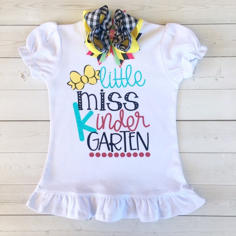 Pu&ograve; includere: Maglietta bianca con orlo a balze, con un fiore giallo, il testo "little miss kindergarten" in lettere colorate e un fiocco a scacchi bianco e nero.