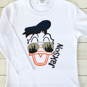 Puede incluir: Camiseta blanca de manga larga con una imagen de dibujos animados de Donald Duck con gafas de sol con un reflejo de castillo. El texto "Jaxson No. 1 Son" est&aacute; impreso en la camiseta.