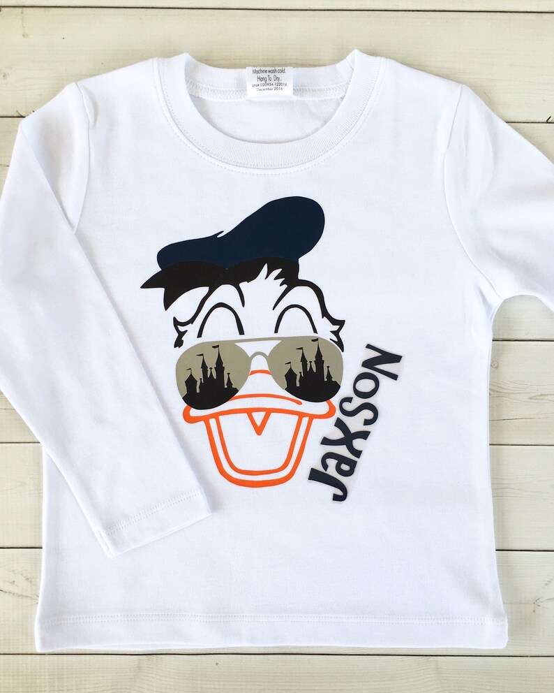 Puede incluir: Camiseta blanca de manga larga con una imagen de dibujos animados de Donald Duck con gafas de sol con un reflejo de castillo. El texto "Jaxson No. 1 Son" est&aacute; impreso en la camiseta.