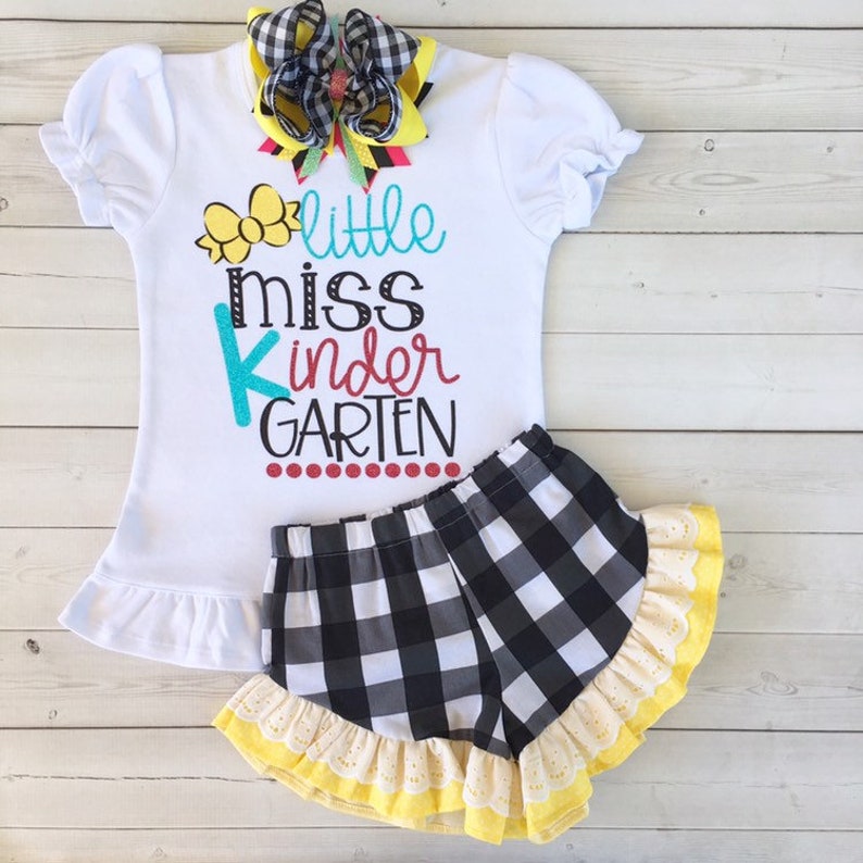 Pu&ograve; includere: Maglietta bianca con gonna a quadri bianchi e neri. La maglietta ha il testo "little miss kindergarten" in lettere colorate.