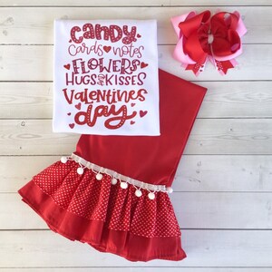 May include: Red and white Valentine's Day outfit for a child. The outfit includes a white t-shirt with the text "candy, cards notes, flowers, hugs & kisses, valentines day" in red, a red skirt with white polka dots and a white ruffle trim, and a red and pink bow.
