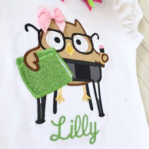 May include: A white t-shirt with an embroidered brown owl wearing glasses and holding a green book. The owl has a pink bow on its head and the name "Lilly" is embroidered in green below the owl.