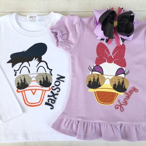 Puede incluir: Dos camisetas blancas y moradas con una imagen de dibujos animados de Donald Duck y Daisy Duck con gafas de sol. El texto "Jackson" y "Kinsley" est&aacute; en las camisetas.