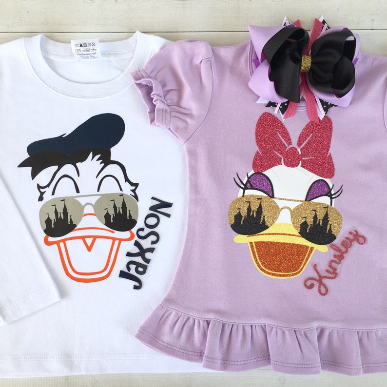Puede incluir: Dos camisetas blancas y moradas con una imagen de dibujos animados de Donald Duck y Daisy Duck con gafas de sol. El texto "Jackson" y "Kinsley" est&aacute; en las camisetas.
