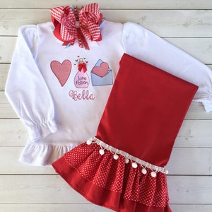May include: White long-sleeve shirt with a red heart, a bottle with the text "Love Potion", and a blue envelope with a red heart. The shirt has a white ruffle at the bottom and the text "Bella" is embroidered below the design. The shirt is paired with red leggings with white polka dots and a white ruffle with white pom-poms at the bottom.
