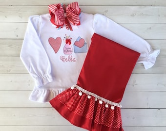 May include: White long-sleeve shirt with a red heart, a bottle with the text "Love Potion", and a blue envelope with a red heart. The shirt has a white ruffle at the bottom and the text "Bella" is embroidered below the design. The shirt is paired with red leggings with white polka dots and a white ruffle with white pom-poms at the bottom.