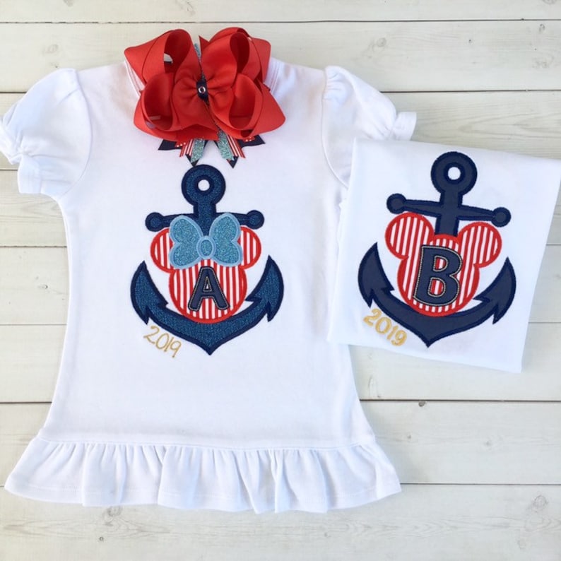 May include: Two white t-shirts with a red, white, and blue anchor design featuring a Mickey Mouse head with a letter on each shirt. The shirts say "2019" on the bottom. The shirt on the left has the letter "A" and the shirt on the right has the letter "B".