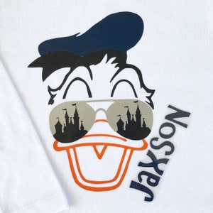Puede incluir: Una camiseta blanca con una imagen de dibujos animados de Donald Duck con gafas de sol con un reflejo de castillo. Las gafas de sol son grises y el castillo es negro. Donald Duck lleva un gorro de marinero azul. El texto "Jaxson" est&aacute; escrito verticalmente en negro en el lado derecho de la imagen.