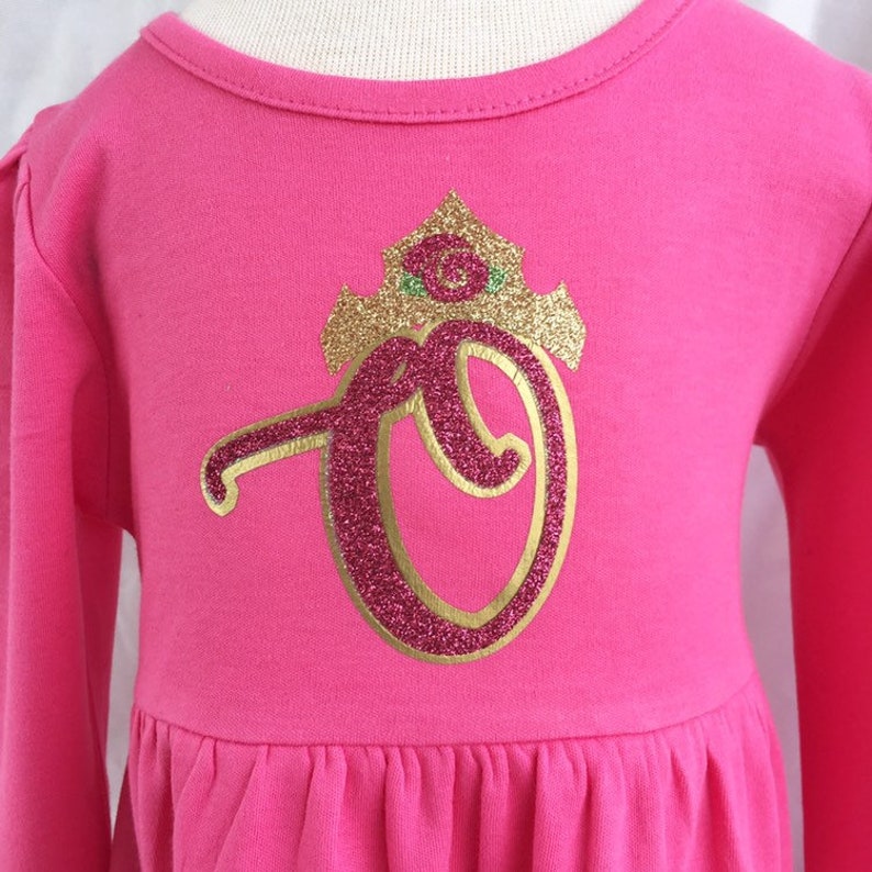 Peut inclure: Une robe rose &agrave; manches longues avec une lettre "O" en paillettes dor&eacute;es et une couronne en paillettes dor&eacute;es surmont&eacute;e d'une rose verte.