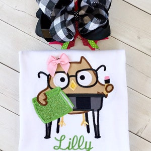 May include: White t-shirt with an embroidered brown owl wearing glasses and holding a green book. The owl is sitting on a chair. The shirt also has the name "Lilly" embroidered in green below the owl.