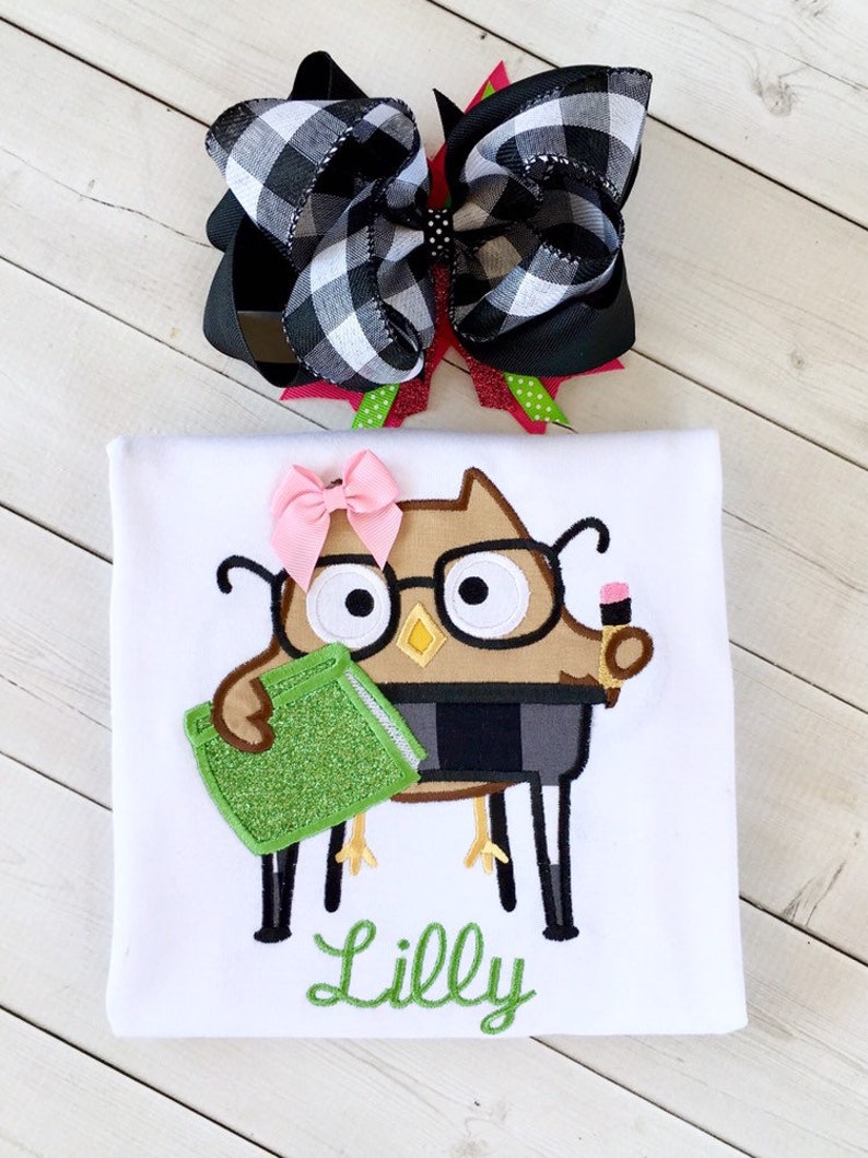 May include: White t-shirt with an embroidered brown owl wearing glasses and holding a green book. The owl is sitting on a chair. The shirt also has the name "Lilly" embroidered in green below the owl.