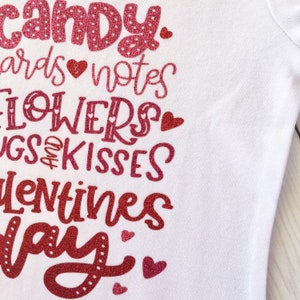 May include: A white t-shirt with a red glitter design that says "Candy, cards, notes, flowers, hugs and kisses, Valentines Day".