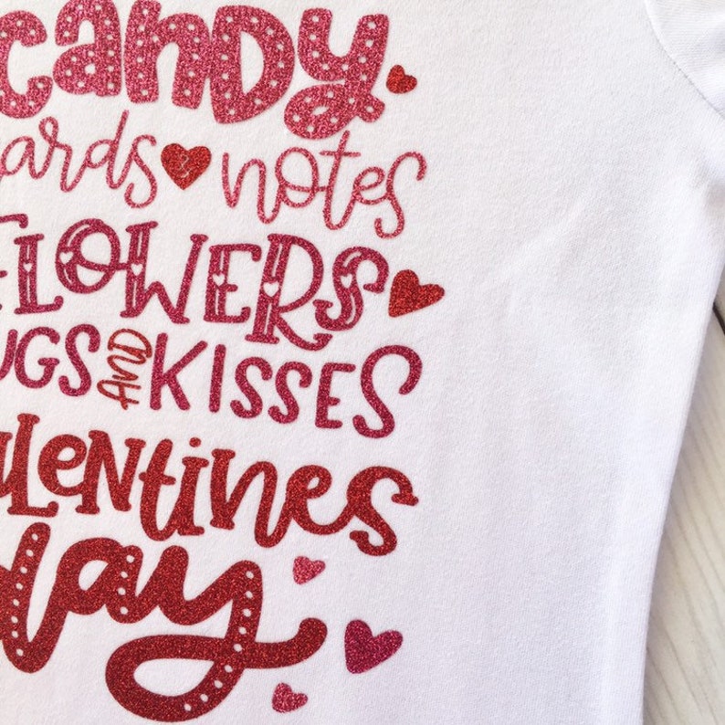 May include: A white t-shirt with a red glitter design that says "Candy, cards, notes, flowers, hugs and kisses, Valentines Day".