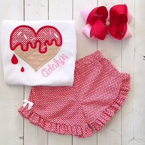 May include: A white t-shirt with a red and pink heart-shaped ice cream cone design and the name "Adelyn" embroidered on the front. The shirt is paired with red and white gingham shorts with a white bow.
