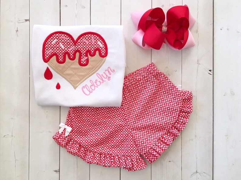 May include: A white t-shirt with a red and pink heart-shaped ice cream cone design and the name "Adelyn" embroidered on the front. The shirt is paired with red and white gingham shorts with a white bow.