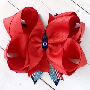 May include: A red and white hair bow with a blue and white circle in the center. The bow is made of grosgrain ribbon and is decorated with glitter.