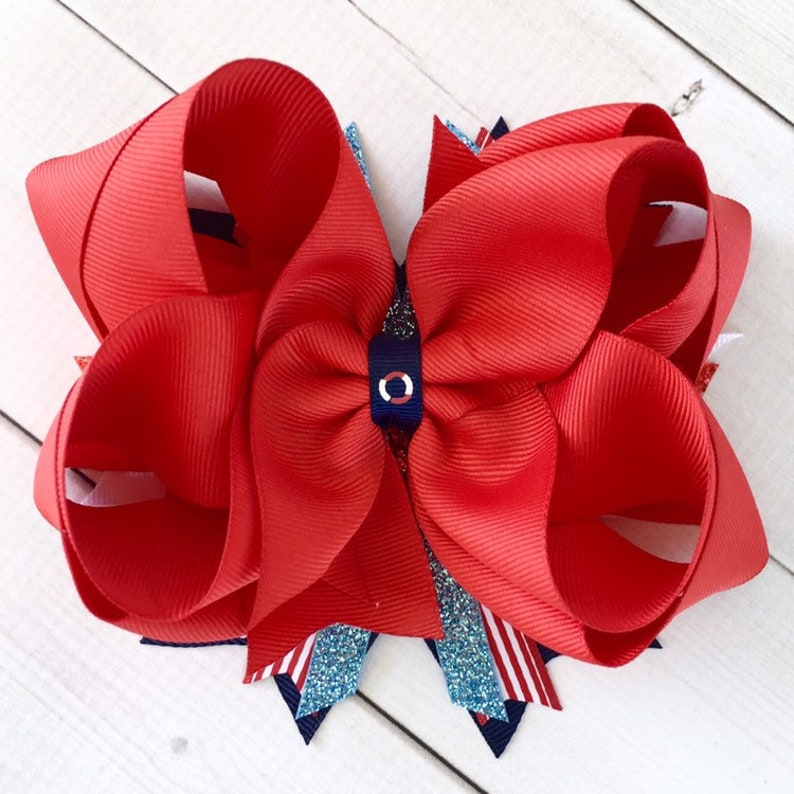 May include: A red and white hair bow with a blue and white circle in the center. The bow is made of grosgrain ribbon and is decorated with glitter.